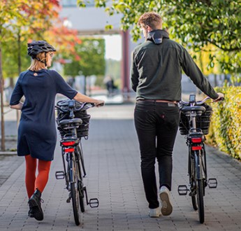 Två cyklister leder sina elcyklar mot universitetets entréhus. 